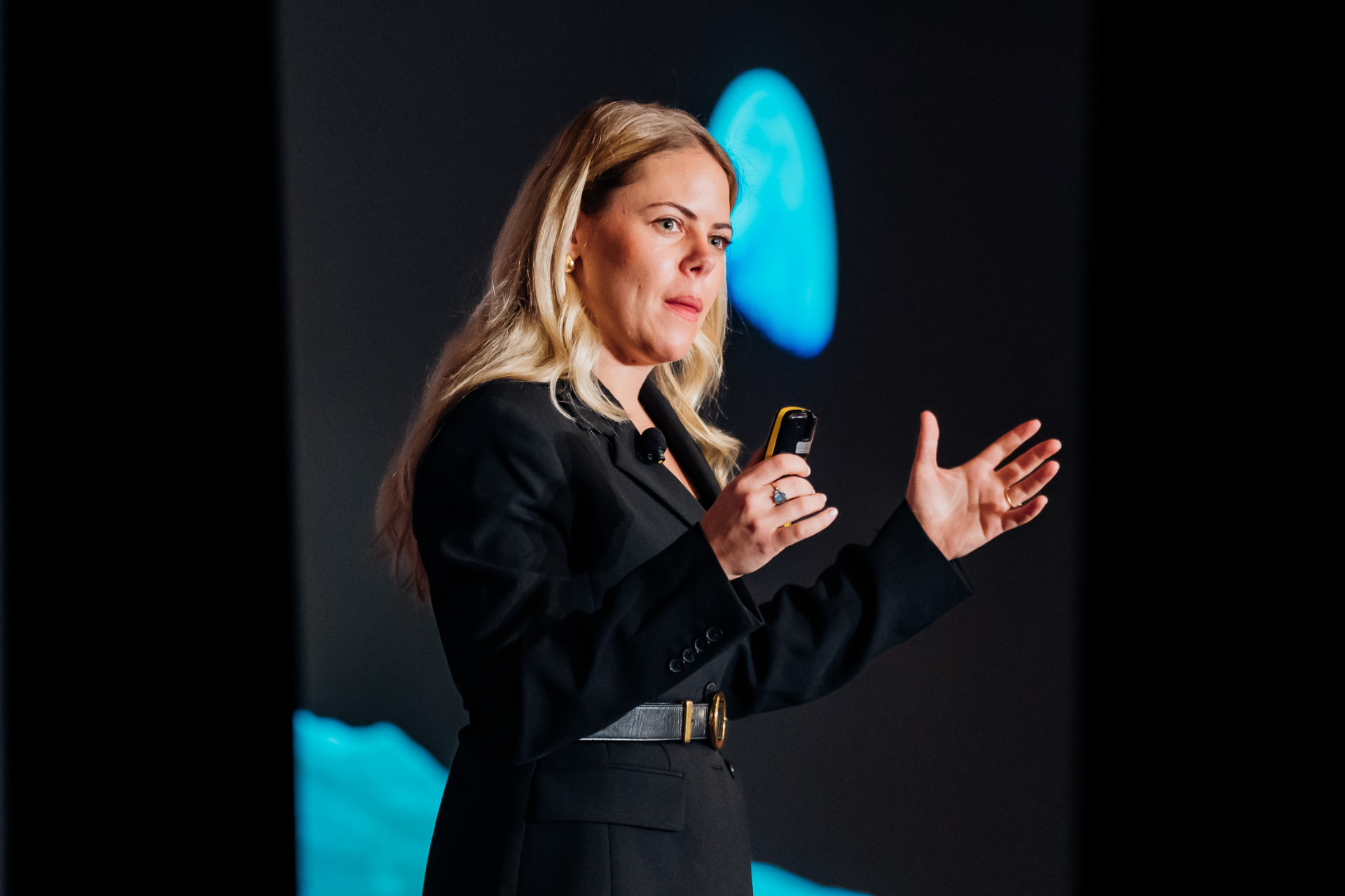 Corporate event photography of a speaker gesturing on stage in front of a backlit corporate logo